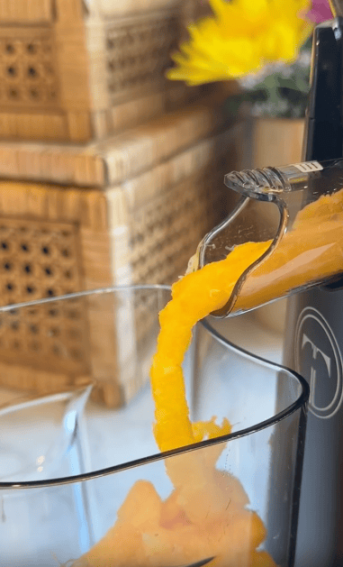 Bright orange juice flows from a juicer spout into a clear glass, showcasing the kitchen appliance's sleek design. Wooden baskets and a yellow flower create a cozy backdrop, highlighting how the juicer complements your counter space.