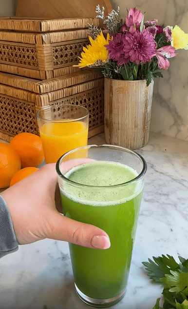 A hand holds a glass of green juice on a marble counter space. Nearby are oranges, a juicer, a glass of orange juice, green herbs, woven containers, and a vase with pink, yellow, and purple flowers.
