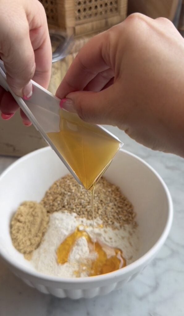 A person pours golden liquid, likely honey, from a small container into a white bowl filled with flour, brown sugar, and oats, with flat folding measuring spoons nearby, preparing ingredients for baking.