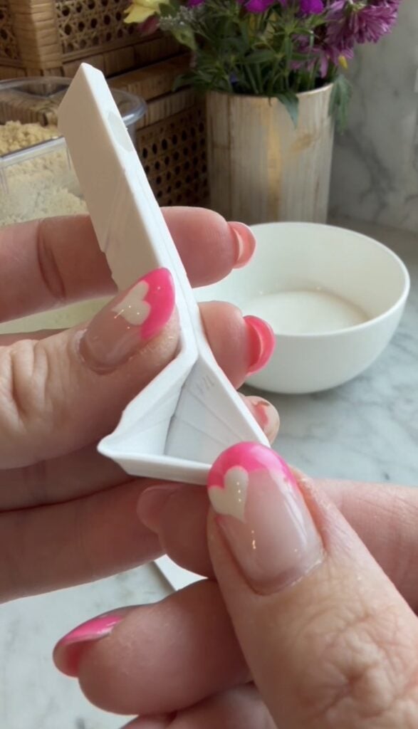A close-up of hands with pink and white heart nail art holding a white plastic razor, with flat folding measuring spoons, a white bowl, and some flowers in the blurred background.