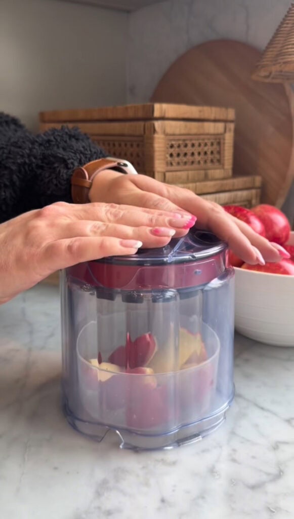 A person presses down on a professional apple slicer containing apple slices, with a bowl of apples and woven baskets in the background on a marble countertop.
