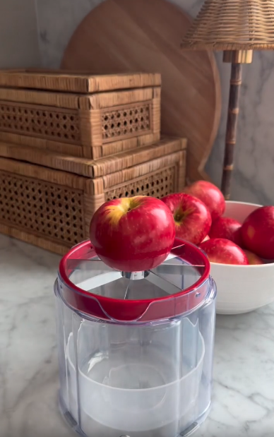 A red apple sits on a clear apple slicer atop a marble countertop. Behind it, a bowl of red apples, two woven baskets, a round wooden cutting board, and a wicker lamp create a professional yet inviting kitchen scene.