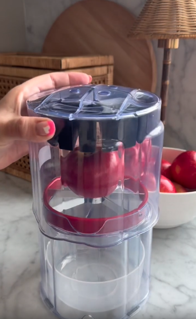 A hand uses a professional Apple slicer, pressing down to slice apples inside a plastic container on a marble countertop. A bowl of apples and kitchen items are visible in the background.