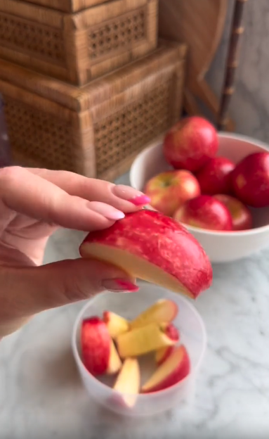 A hand with pink nails holds a red apple slice above a container with more neatly cut slices, showing the precision of a professional apple slicer. In the background, a white bowl of whole apples and wicker baskets are visible.
