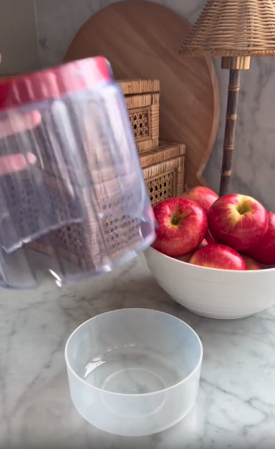 A person holds a clear plastic container with a red lid above a white bowl filled with sliced apples. Wicker baskets and an apple slicer sit on the marble surface, giving the scene a professional kitchen feel.