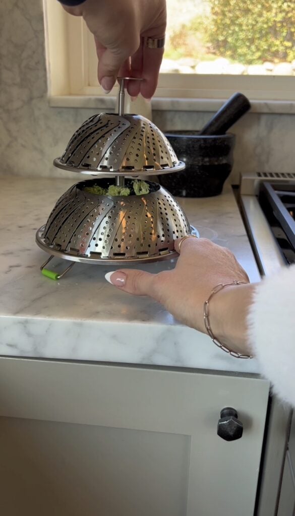 A hand holds two stacked metal vegetable steamers on a marble kitchen counter, with a mortar and pestle and a stovetop in the background.