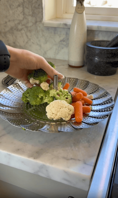 A hand places broccoli, cauliflower, and baby carrots onto a metal steamer basket on a kitchen counter near a mortar and pestle.