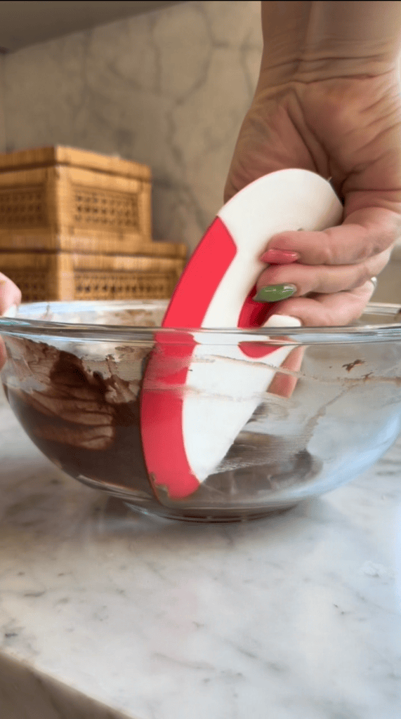 A hand with green-painted nails uses a red and white spatula to mix chocolate batter in a clear glass bowl on a marble countertop. Wicker boxes are visible in the background.