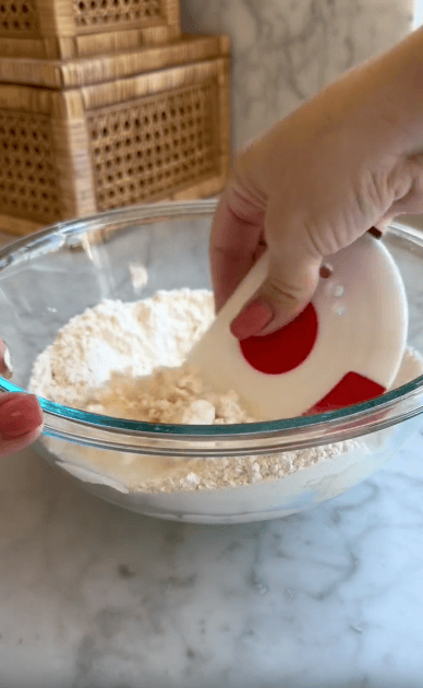 A hand uses a white and red dough scraper to mix flour in a clear glass bowl on a marble countertop. Woven wooden boxes are stacked in the background.