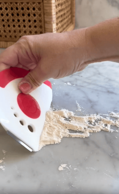 A hand uses a red and white dough scraper to gather flour and dough remnants on a marble countertop.