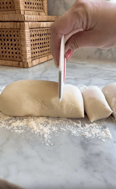 A hand uses a white dough scraper to cut a log of dough on a floured marble countertop, with wicker containers in the background.