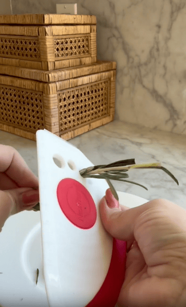 A person uses a white and red plastic kitchen tool to strip leaves from a sprig of rosemary. Two woven storage boxes sit on a marble countertop in the background.