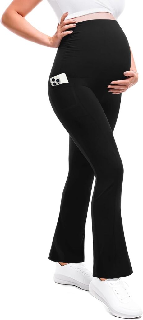 A pregnant person wearing black maternity pants with a phone in the side pocket, a white top, and white sneakers, with one hand on their belly.