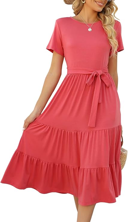 Woman wearing a short-sleeved, coral pink, tiered midi dress with a waist tie, holding the skirt with one hand. She has wavy hair and accessorizes with a necklace and a woven bag.