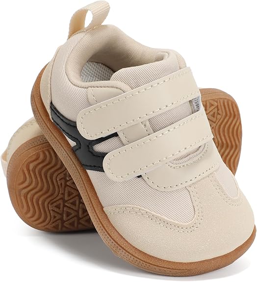 A pair of beige toddler sneakers with brown rubber soles, double Velcro straps, and a black accent on the side, displayed with one shoe slightly leaning on the other.