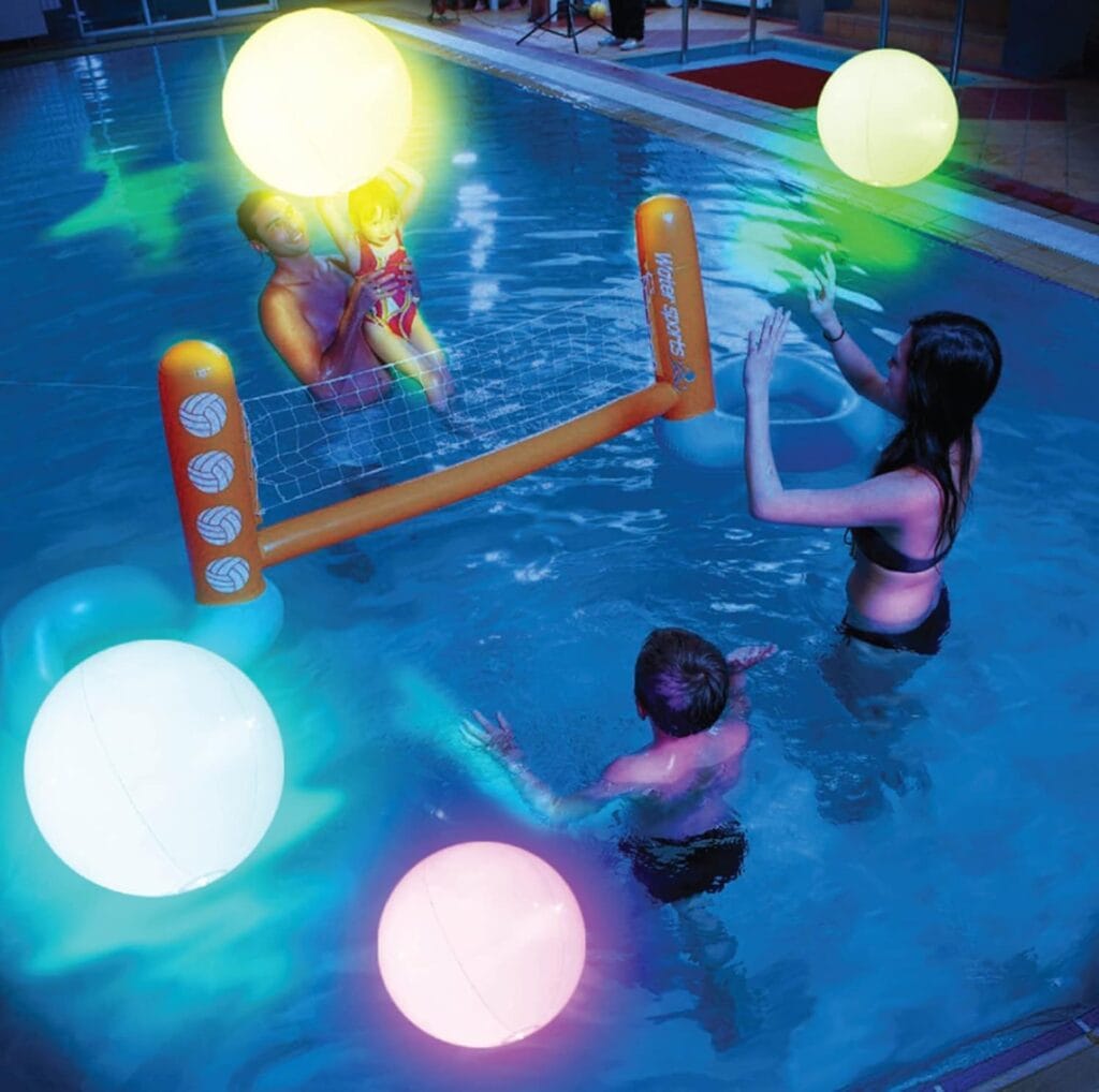 A family plays volleyball in a swimming pool at night with glowing, colorful beach balls. An adult holds a child on one side of the net, while another adult and a child stand on the other side.