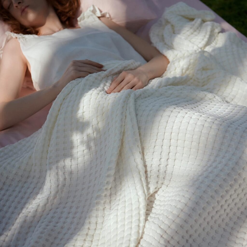 A person lies on a bed covered with a textured white blanket. Soft sunlight casts shadows on the blanket, and the person is wearing a light sleeveless top, partially visible from shoulders to upper legs.