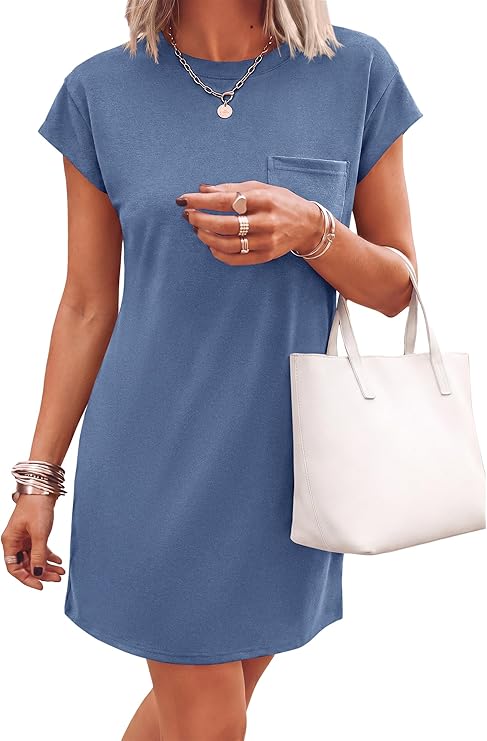 A woman wearing a short-sleeve, blue t-shirt dress with a chest pocket, holding a white tote bag and accessorized with layered necklaces, rings, and bracelets.