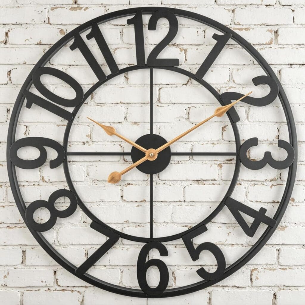 Large black metal wall clock with open frame and bold numbers, featuring gold clock hands, mounted on a white painted brick wall. The time shown is approximately 2:10.