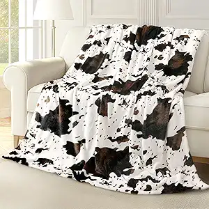 A white sofa is draped with a cozy blanket featuring a black and brown cow print pattern. The room has light walls and a large window letting in natural light.