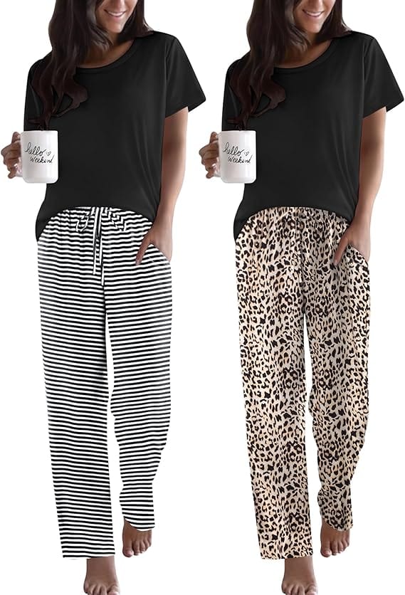 Two women stand barefoot, each holding a white mug. One wears a black t-shirt with black-and-white striped pants, the other wears a black t-shirt with beige leopard-print pants. Both are smiling.