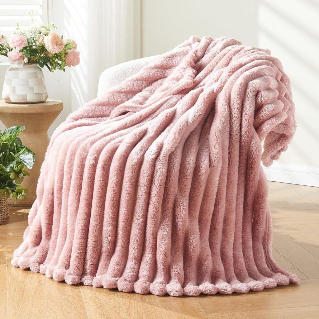 A plush, light pink faux fur blanket draped over a white chair in a sunlit room with wooden floors, near a small table with a vase of flowers.