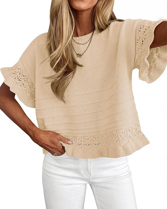 Woman wearing a beige short-sleeve knitted top with lace details on the sleeves and hem, paired with white pants. She has long, wavy blonde hair and is accessorized with layered necklaces.