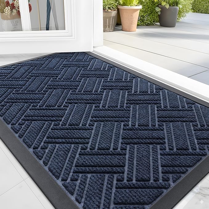 A dark blue textured doormat with a geometric woven pattern lies in front of an open white door, leading to a bright patio with potted plants—perfect for upgrading your entryway with Monday Deals.