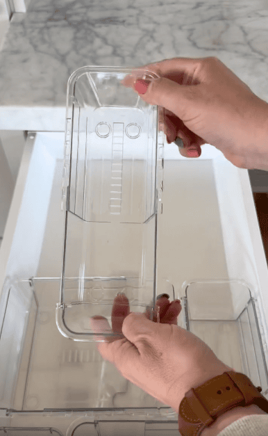 A person holds a clear plastic organizer tray above a drawer with similar trays, displaying its size and shape; a marble countertop is visible in the background.