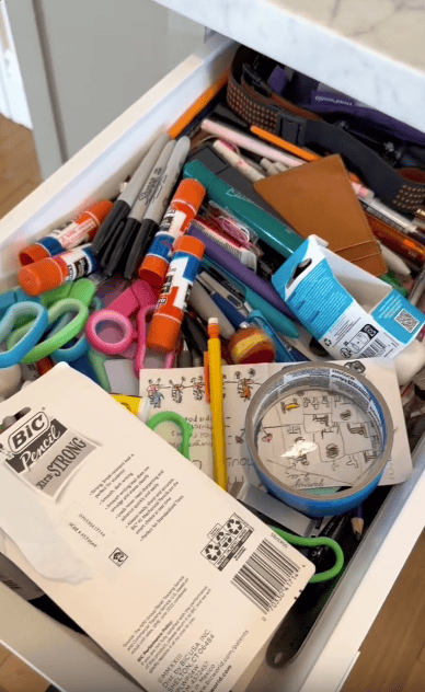 An open drawer filled with assorted school and office supplies, including glue sticks, scissors, pens, markers, tape, a pencil, and various packaging and papers in a disorganized manner.