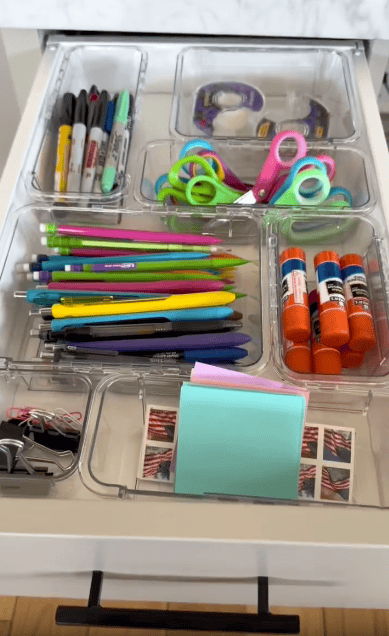 An organized drawer with clear dividers holding markers, scissors, tape, pens, pencils, glue sticks, binder clips, sticky notes, and stamps, neatly arranged for easy access.