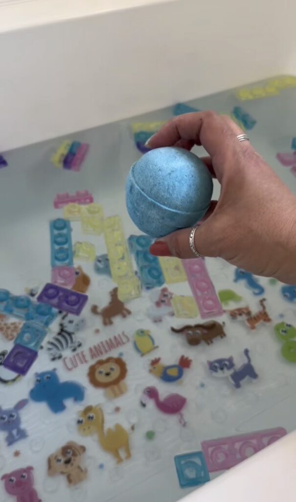 A hand holds a blue bath bomb over a bathtub filled with water, floating foam blocks, and colorful animal-themed bath stickers, including lions, flamingos, and cats.