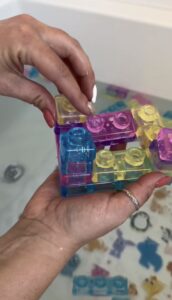 A person holds colorful translucent toy blocks, including blue, yellow, and pink pieces, above a bathtub filled with water and more floating blocks.
