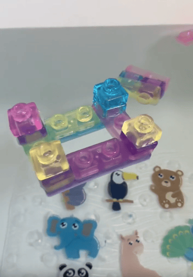 Colorful translucent toy building blocks are stacked in a small structure above animal stickers, including an elephant, bear, panda, toucan, alpaca, and peacock, on a smooth white surface.