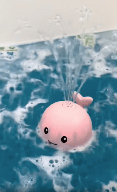 A pink whale-shaped bath toy floats in blue water, spraying water upwards from a hole on top of its head, surrounded by bubbles.