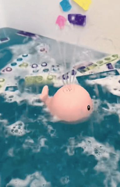 A pink, whale-shaped toy floats in bubbly blue water, spraying water upward. Colorful foam bath letters and numbers are stuck on the wall in the background.
