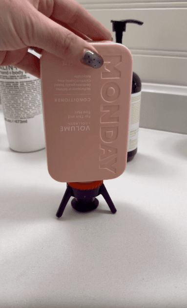 A hand holds an upside-down pink MONDAY brand conditioner bottle on a purple bottle holder on a white bathroom counter. Other bottles are visible in the background.