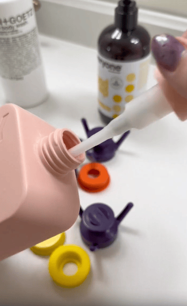 A hand dips a white stick into a pink bottle. In the background are small colorful watering can-shaped objects and bottles of toiletries on a white surface.