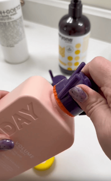 A hand with purple, speckled nails unscrews the purple cap of a peach-colored bottle labeled "DAY." Other bottles and containers are visible in the blurred background on a countertop.