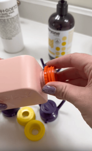 A person with painted nails attaches an orange plastic pump to a pink bottle, with other bottles and colorful pump tops placed on a white countertop in the background.