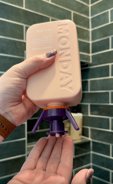 A hand squeezes an upside-down pink MONDAY brand conditioner bottle with a purple cap, dispensing product onto the other hand in a tiled bathroom.