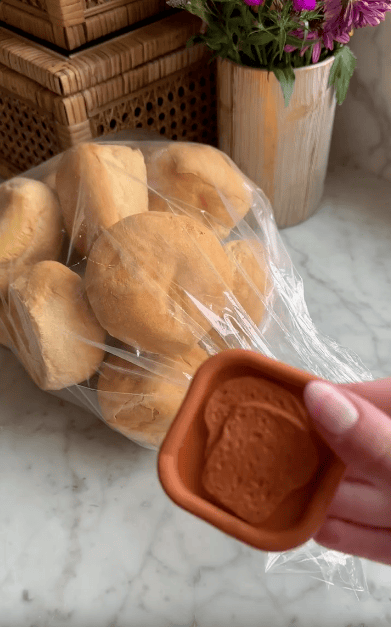 A hand holds a small terracotta dish with a bread design in front of a plastic bag filled with round bread rolls on a marble countertop, with baskets and flowers in the background.