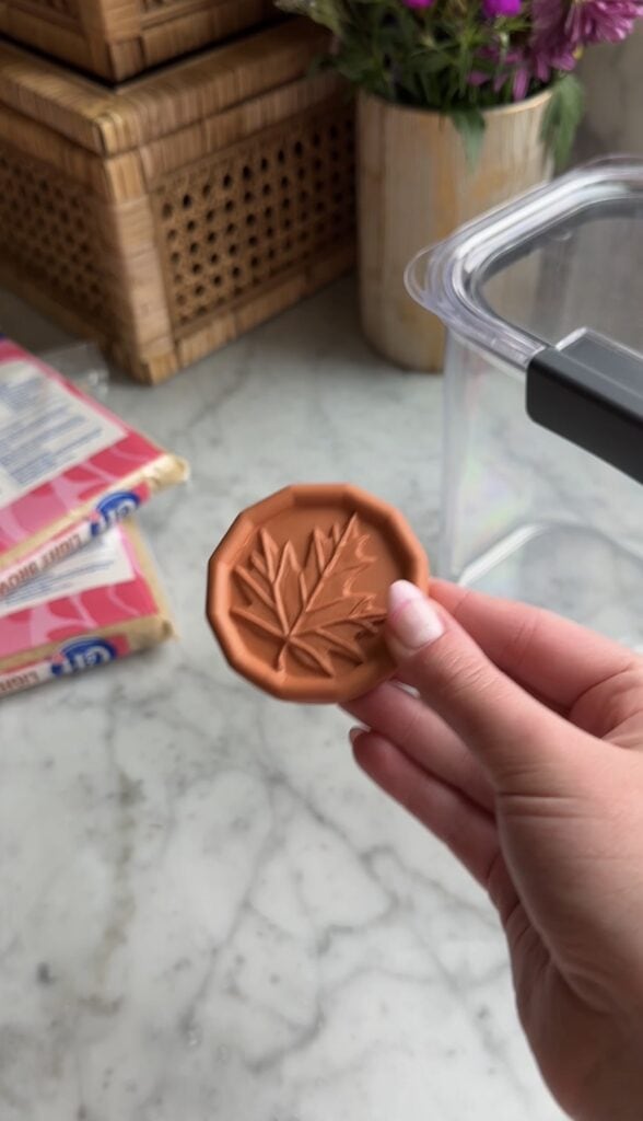 A hand holds a round terra cotta disk with a leaf design. In the background, there are packages of sugar, a clear container, and a woven basket on a marble countertop.