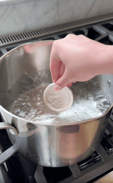A hand holds a round white object above a pot of boiling water on a stove, about to place it into the water.