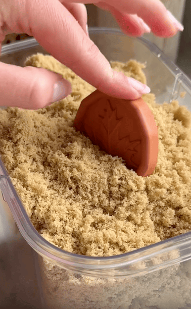 A hand presses a brown terracotta disc with a leaf design into a container filled with light brown sugar.