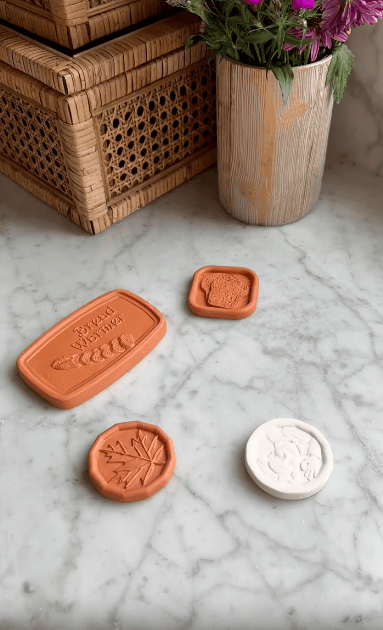 Four clay stamps with various designs, including leaves and bread, are displayed on a marble surface. Behind them are woven baskets and a vase with purple flowers.