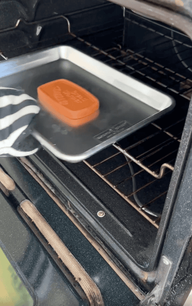 A gloved hand places a metal baking tray with a rectangular terracotta bread warmer into an open oven.
