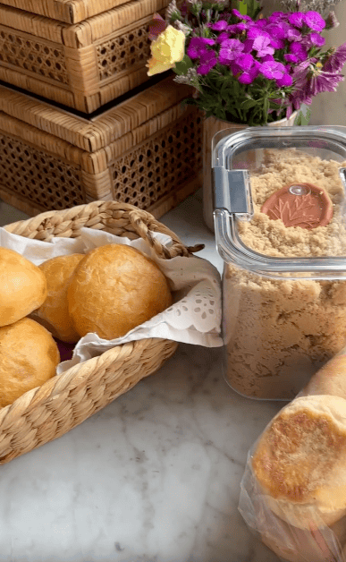 A woven basket filled with round bread rolls sits on a marble countertop next to a container of brown sugar with a scoop inside. Behind them are wicker boxes and a vase of colorful flowers.