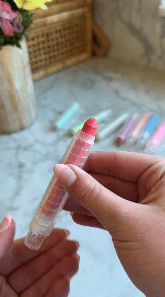 A hand holds a clear plastic tube with a pink lipstick or crayon inside. Several similar tubes in various colors are blurred in the background on a marble surface.