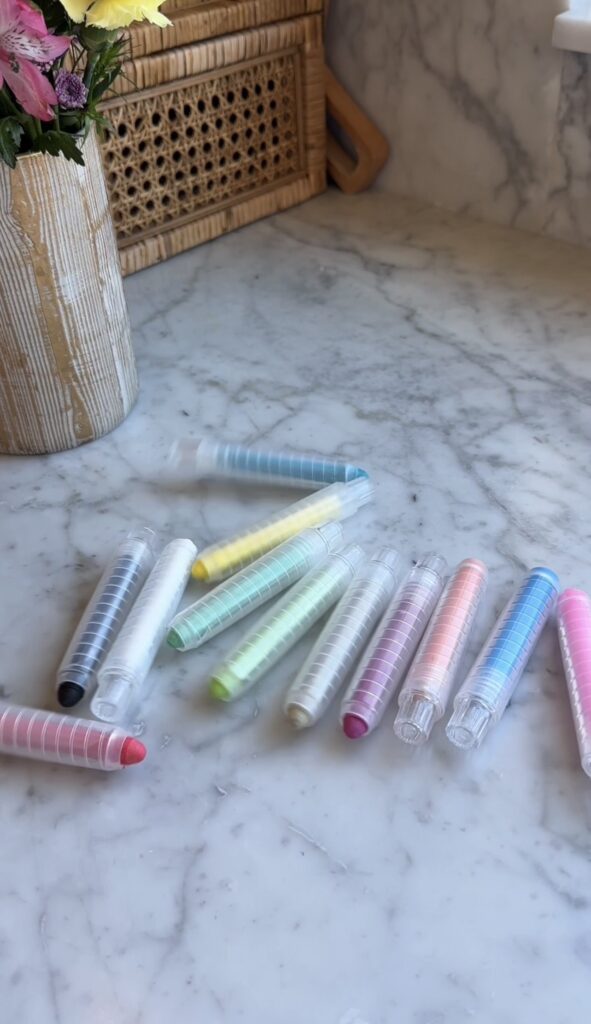 Plastic pastel-colored markers with clear caps are scattered on a marble surface near a wooden container. The markers include shades of pink, blue, green, yellow, gray, and black.